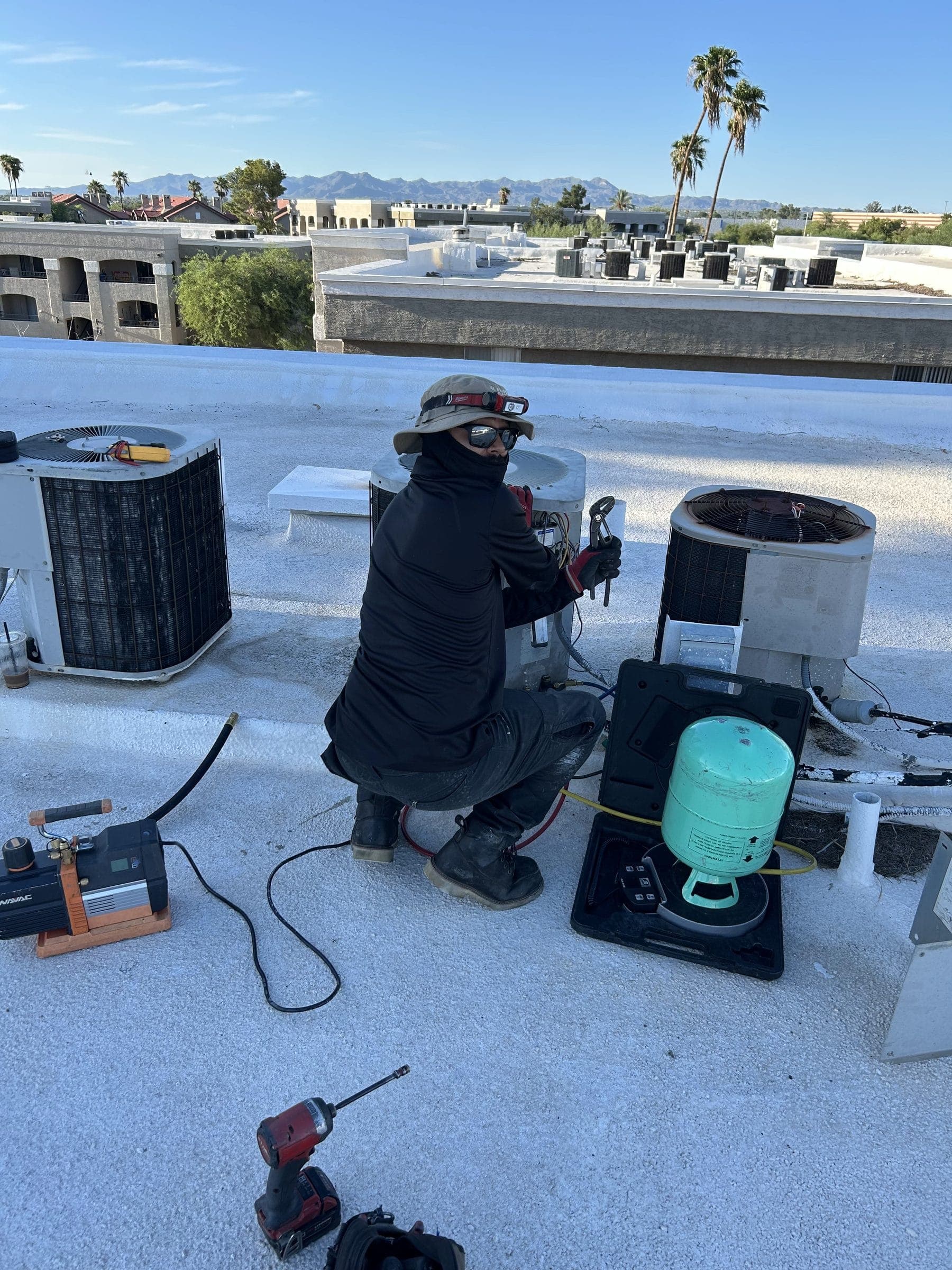 Technician servicing a rooftop condenser with gauges and a refrigerant cylinder
